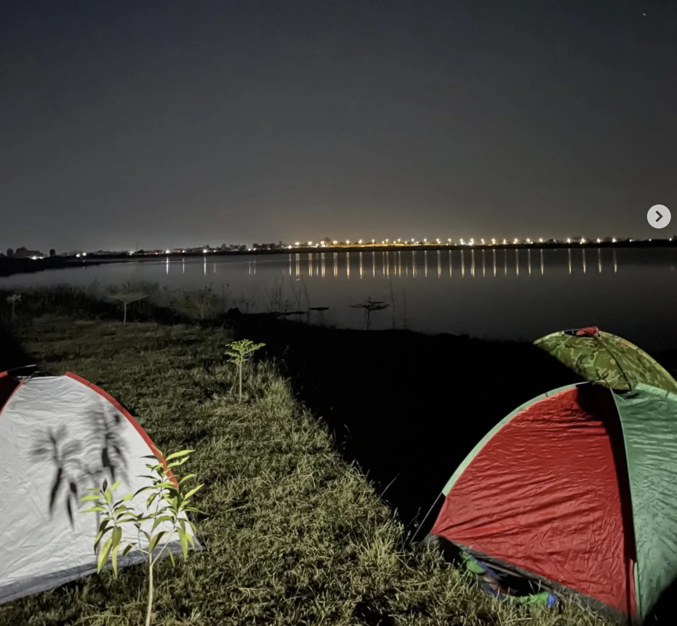 Tents pitched by a waterfront at night with city lights reflecting on the water