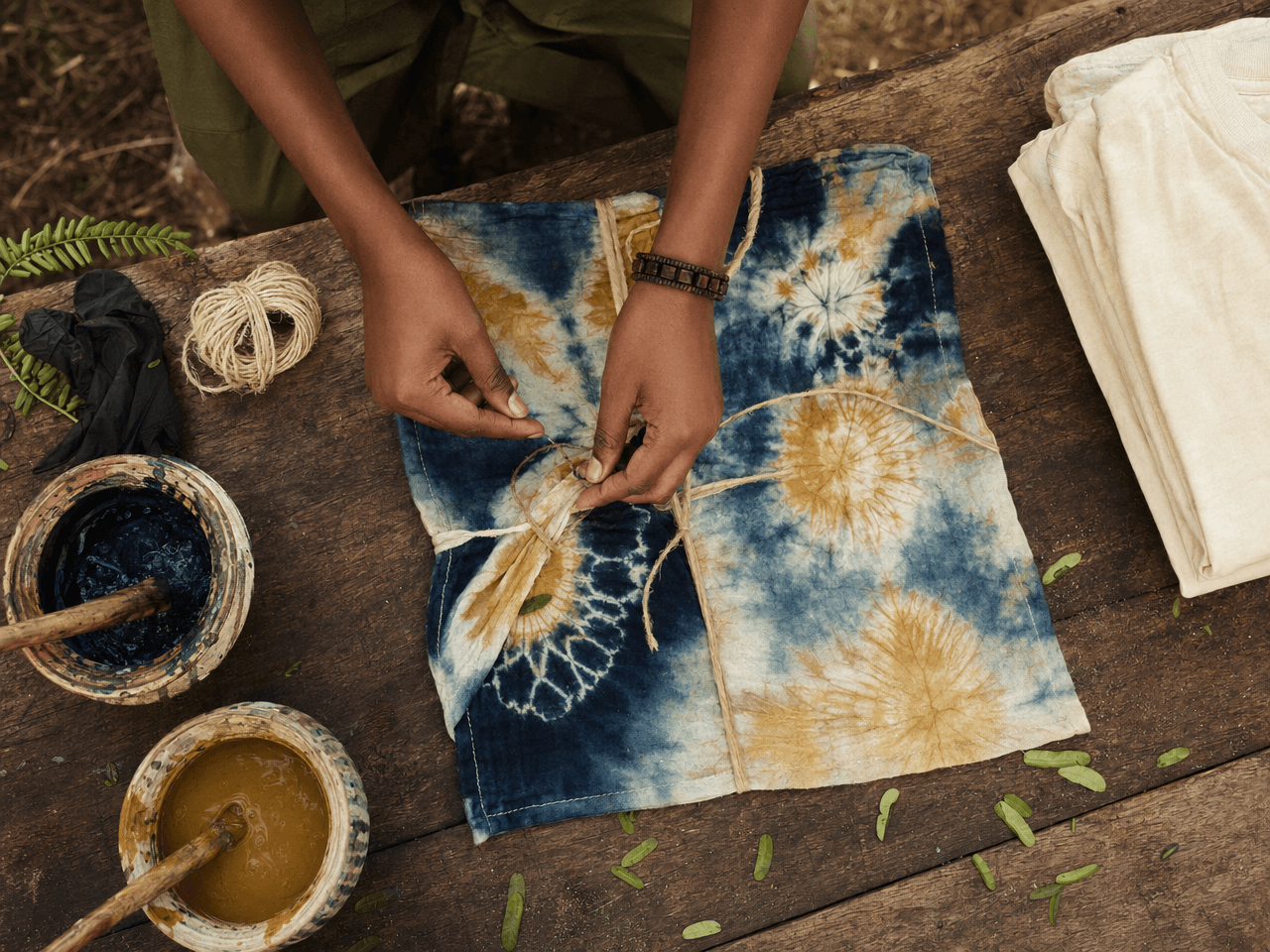 Top-down view of hands tie-dyeing an indigo and gold Adire bandana on a wooden trestle table